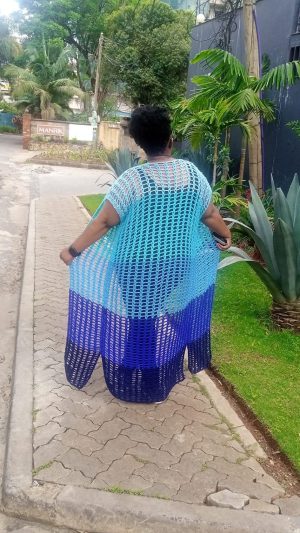 A woman wearing a long, ombré crochet cover-up in shades of light blue to deep navy, walking on a paved path surrounded by greenery. The cover-up has an open mesh design.